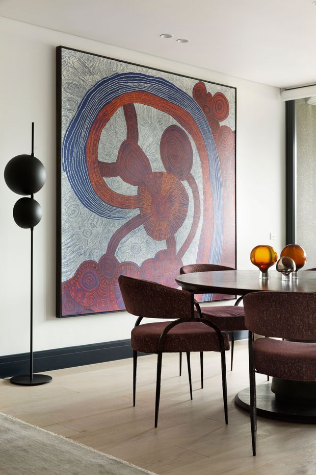 A refined dining area with a dark round dining table and upholstered burgundy dining chairs, set against white walls with dark skirting. A large framed abstract artwork provides a bold red, navy, and grey focal point, complemented by a sculptural black floor lamp and amber glass objects on the table. Pale timber flooring and soft neutral furnishings create a polished, contemporary atmosphere.