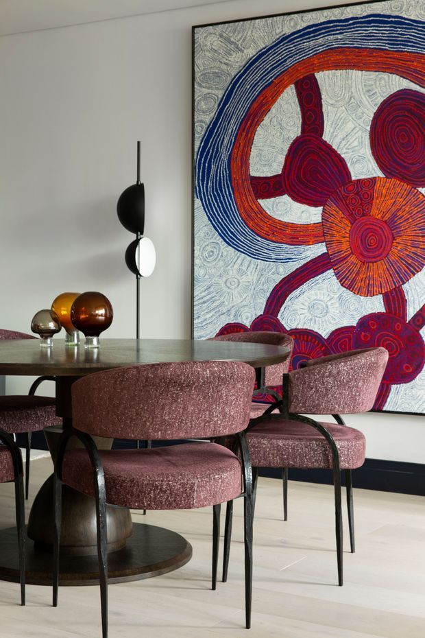 A refined dining area with a round dark stone or wood-topped table, upholstered dusty-rose dining chairs, and sculptural pendant lighting. A large colourful abstract artwork with concentric circular motifs dominates the wall, complemented by small decorative glass objects on the table. The room has pale flooring, white walls, and a dark skirting board, creating a polished, eclectic atmosphere.