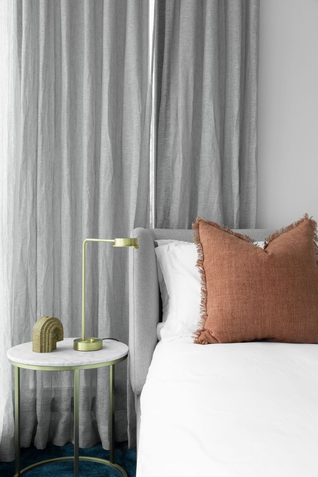 A styled bedroom vignette featuring a white bed with a light grey upholstered headboard, a large rust-brown fringed accent cushion, full-height sheer grey curtains, and a round marble-topped side table with slender brass-toned legs. A slim brass desk lamp and small sculptural object sit on the table. The overall look is refined, contemporary, and softly layered with neutral tones and a warm terracotta accent.