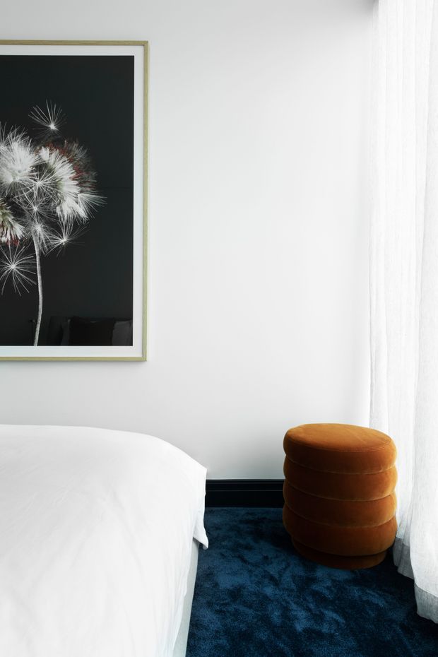 A bright, refined bedroom corner with a white bed, white walls, sheer floor-length curtains, deep navy carpet, and a rounded burnt-orange upholstered stool. A large framed black-and-white botanical photograph provides a graphic feature accent.
