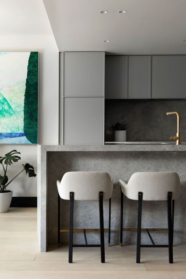 Contemporary kitchen interior featuring a compact grey island with an integrated stone-look benchtop and waterfall-style side panel, matching grey full-height cabinetry, dark textured splashback, and a brass gooseneck tap. Two upholstered bar stools sit at the island. Pale timber flooring, white walls, recessed ceiling lights, a potted plant, and colourful artwork complete the refined neutral space.