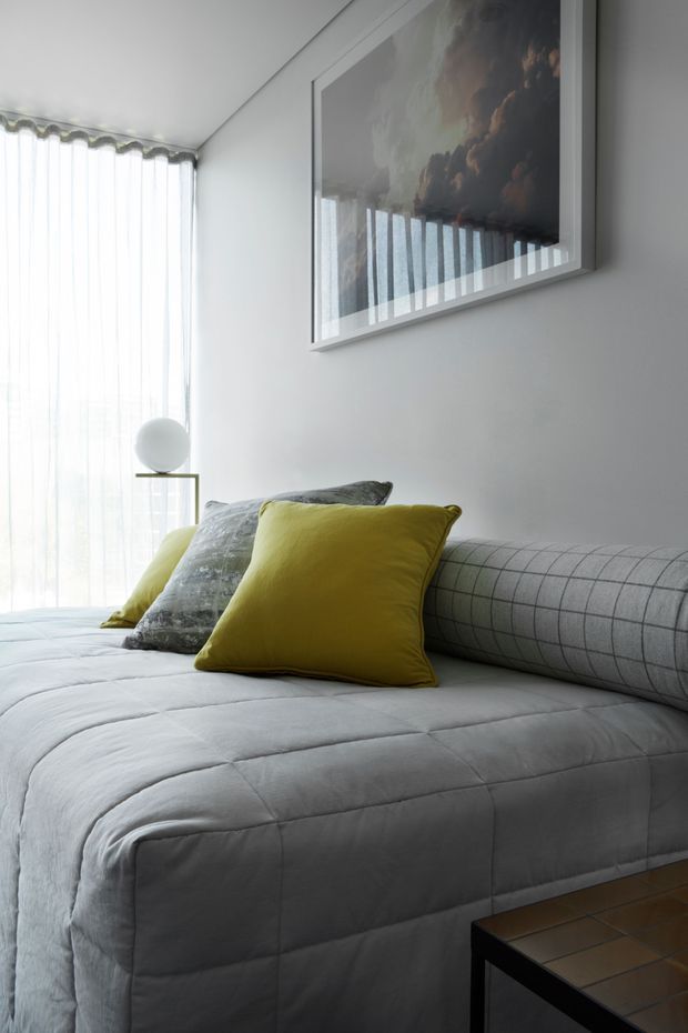 A bright, compact bedroom with a neatly made upholstered bed, pale grey quilted bedding, a grey checked cylindrical bolster, and contrasting mustard-yellow and muted patterned cushions. Floor-to-ceiling sheer curtains filter daylight beside a small minimalist side table with a spherical lamp. A large framed artwork hangs above the bed, and a dark wood or black-framed tiled side table is visible in the foreground.