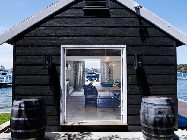 Front-on exterior view of a small black weatherboard-clad gable-roofed structure beside a body of water. A central glazed doorway reveals a compact furnished dining area inside, while two timber barrels flank the entrance. The building has white roof and door trims, dark horizontal cladding, and a marina-like waterfront setting.