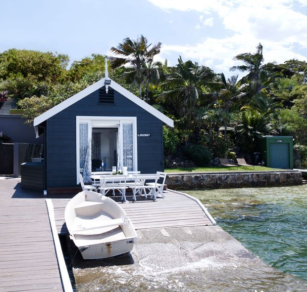 A compact dark-painted timber-clad waterside cottage with a white gabled facade, opening onto a timber jetty and concrete boat ramp. Outdoor dining furniture and a small boat sit beside the water, with dense tropical planting surrounding the property.
