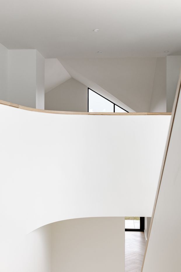 Minimalist interior view of a sculptural curved stairwell or landing, dominated by smooth white walls and a sweeping balustrade. A light timber handrail traces the curve, while a sharply angled black-framed window and glimpses of pale timber flooring add contrast. Recessed ceiling lights and strong geometric forms create a bright, refined architectural composition.
