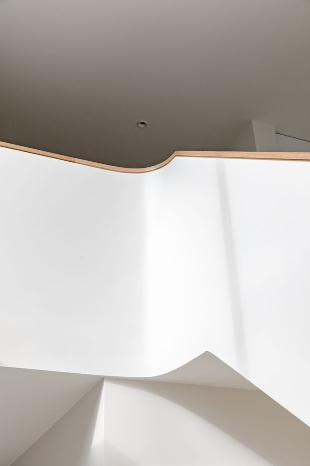 Abstract architectural interior view focused on a sculptural stairwell or ceiling transition. Smooth white plaster surfaces form angular and curved planes, with a dark grey ceiling edged by a narrow light timber trim. Minimal recessed lighting and strong natural light emphasize the geometric composition; no furniture is visible.