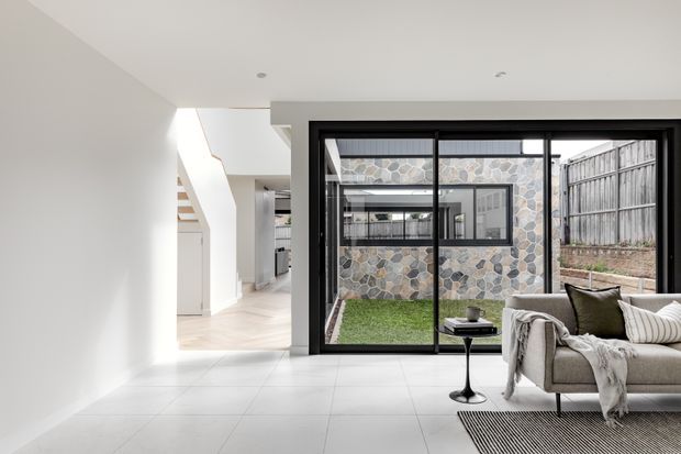 Bright contemporary open-plan living area with a minimalist white interior, pale tiled flooring, and large black-framed sliding glass doors opening to a small landscaped courtyard. A neutral upholstered sofa with cushions and a throw occupies the right side, while a stairway and kitchen circulation area are visible in the background. The courtyard features a distinctive irregular stone-clad wall and timber boundary fencing.