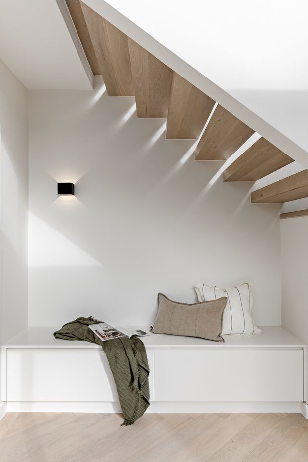 Minimalist interior beneath a timber-lined stairway, featuring a built-in white window seat or storage bench with neutral cushions, soft wall lighting, and pale herringbone timber flooring. The open-riser stair structure creates a strong architectural feature overhead, with a calm, restrained palette of white, beige, and light wood.