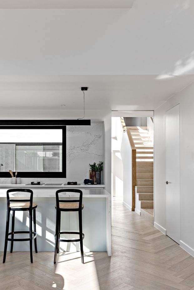 Bright contemporary open-plan kitchen with a white island and black-framed window, paired with two black bar stools. A marble-look splashback, pale herringbone timber flooring, white walls, and a timber staircase create a clean, light-filled interior. The view also includes a hallway and stairway leading deeper into the home.