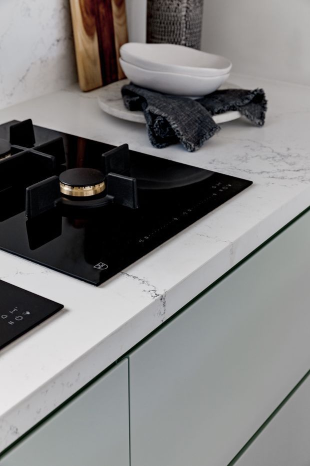 Close-up detail of a refined contemporary kitchen, focusing on a black glass gas cooktop set into a white veined stone benchtop. Pale grey-green flat-panel cabinetry with discreet integrated handles fills the lower portion, while a white vessel and dark textile sit on a tray in the background. The composition is minimal and monochromatic with subtle brass detailing on the cooktop burner.