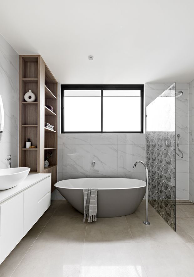 A bright contemporary bathroom featuring a freestanding oval bathtub, wall-mounted white vanity with vessel basin, open timber shelving, large-format marble-look wall tiles, grey floor tiles, and a clear glass shower screen with patterned feature tiles. Black-framed awning windows provide strong contrast against the predominantly white and grey finishes.