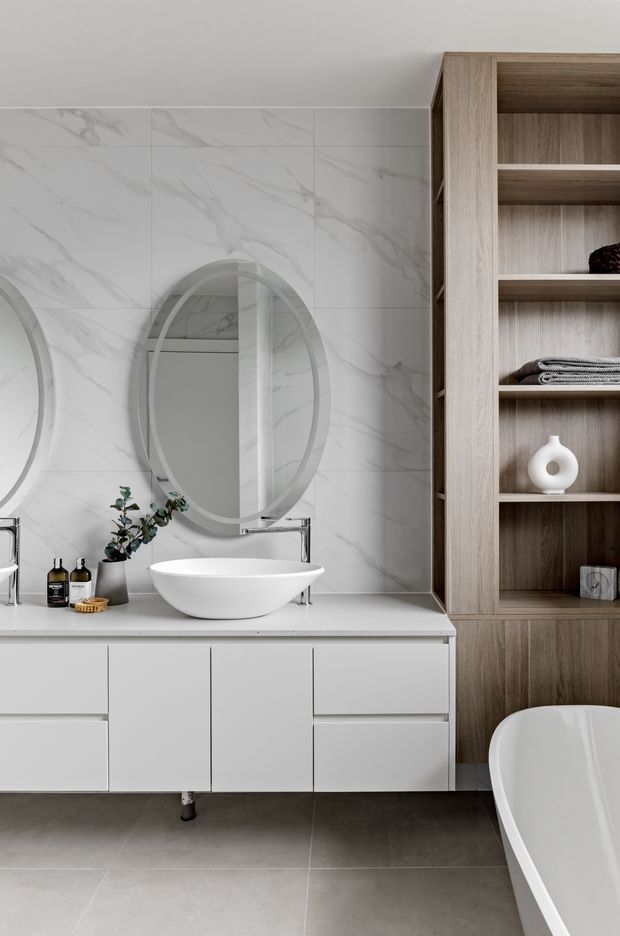 A refined contemporary bathroom featuring a floating white vanity with integrated drawers, a white vessel basin, wall-mounted chrome tapware, twin oval mirrors, and a large-format white marble-look tiled wall. A full-height light timber open shelving unit provides storage for towels and accessories, while part of a freestanding white bathtub is visible at the right. The floor is finished in large grey tiles, creating a calm, neutral palette with warm timber contrast.