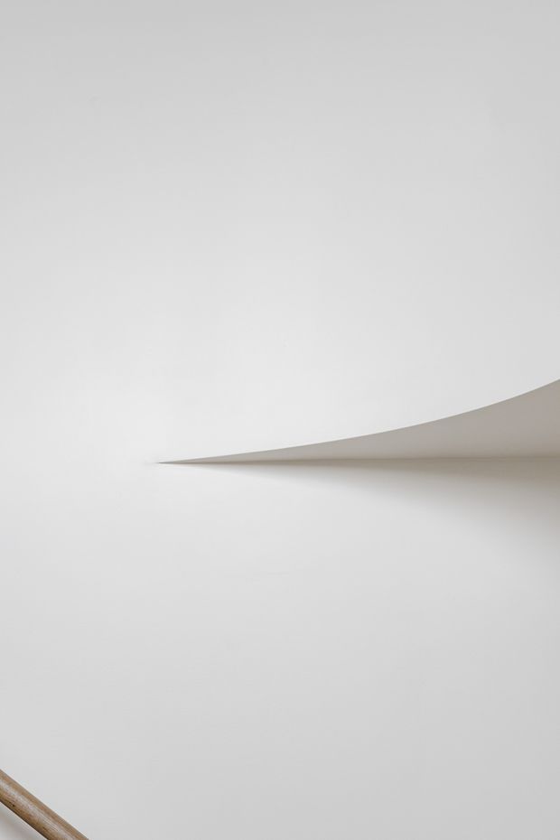 Minimalist architectural detail showing a smooth white curved or folded ceiling/wall feature projecting into the space, with soft shadowing and a small glimpse of a timber handrail at the lower left. The image is abstract, monochromatic, and focused on sculptural form and indirect light.