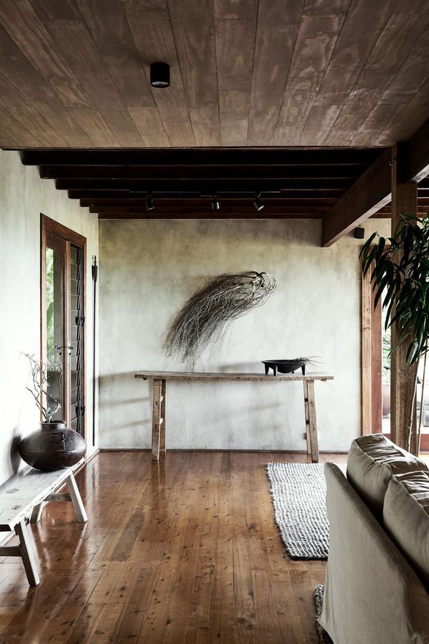 A warm, rustic living room with dark timber floorboards, a timber-lined ceiling and exposed dark beams. A long timber console table sits against a softly textured plaster wall beneath an organic wall sculpture, with a neutral sofa, woven rug, ceramic vessel, and abundant natural light from tall timber-framed glazing. The room has a restrained, earthy and handcrafted character.