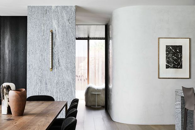 A refined open-plan dining and living area with a long timber dining table and dark upholstered chairs in the foreground. A dramatic grey-veined stone-clad feature wall incorporates a vertical brass handle, while dark timber cabinetry, pale flooring, a curved white plaster wall, and a glimpse of a lounge area create a sophisticated, sculptural interior. Framed monochrome artwork and a black-framed glazed opening add contrast.