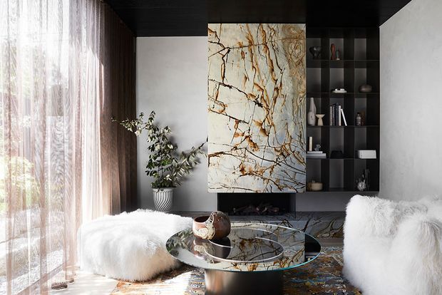 A refined contemporary living room with a dark ceiling and built-in black shelving surrounding a dramatic veined stone feature panel and low media unit. A round glass coffee table, sculptural seating upholstered in white faux fur, sheer floor-to-ceiling curtains, a potted plant, and a patterned rug create a luxurious, high-contrast interior.