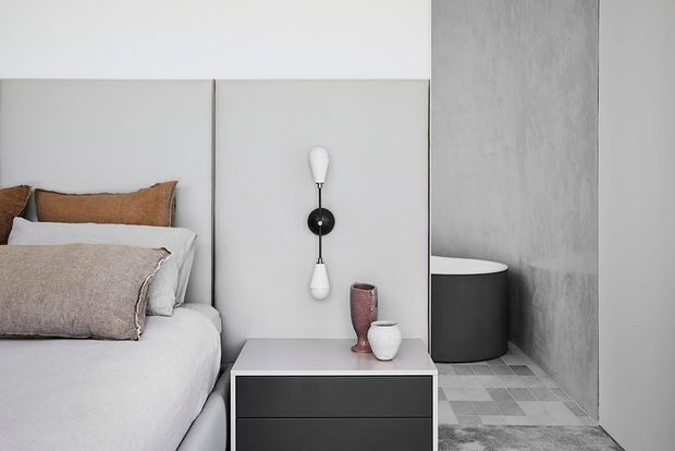A close-up view of a minimalist bedroom corner featuring a partially visible bed with white bedding and neutral textured pillows, a tall pale upholstered or paneled headboard, a black-and-white wall sconce, and a compact dark bedside table with ceramic vessels. The room has a grey textured wall and a geometric grey tiled floor, creating a restrained contemporary interior.
