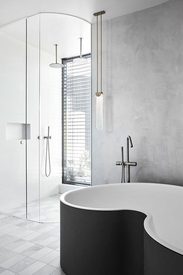 A bright contemporary bathroom featuring a sculptural freestanding white bathtub with a dark exterior, a frameless glass walk-in shower, wall-mounted rain showerheads, a textured grey concrete-look wall, pale geometric floor tiles, and dark minimalist tapware. A tall louvred window provides natural light.