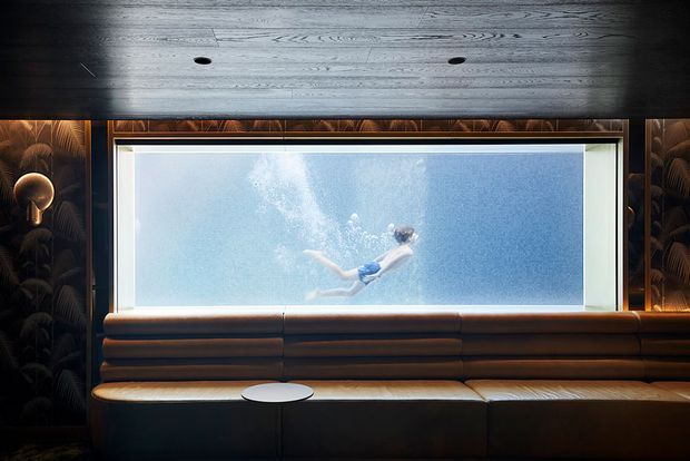 Moody, luxurious interior featuring a long built-in brown leather banquette beneath a large underwater viewing window. A swimmer is visible in the blue pool beyond the glass. Dark timber ceiling panels, recessed spotlights, and a dramatic dark botanical wall covering frame the window, with a small round table in front of the seating.