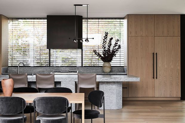 A refined open-plan kitchen and dining area with a long stone island and raised dining counter, integrated timber cabinetry, black appliances, large horizontal windows with blinds, and a contemporary dining table with upholstered chairs. A dark sculptural pendant light and a ceramic vase provide focal accents. The room has a warm, sophisticated palette of timber, charcoal, grey stone, and black.