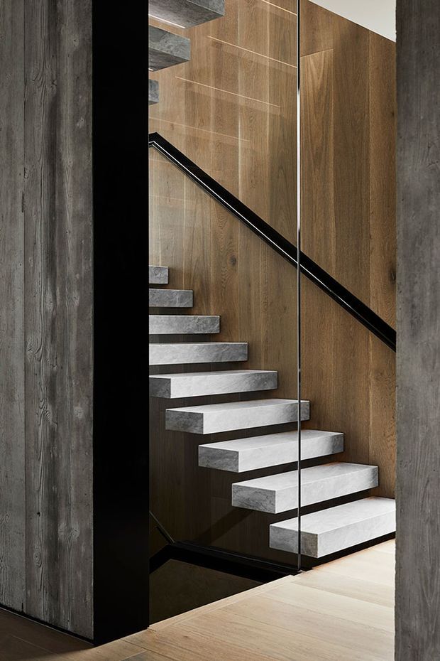 A refined modern stairway viewed through a glass balustrade, with floating pale concrete or stone treads set against a warm timber-panelled wall. Dark structural framing, a black handrail, and charcoal textured wall surfaces create a strong linear, high-contrast composition.