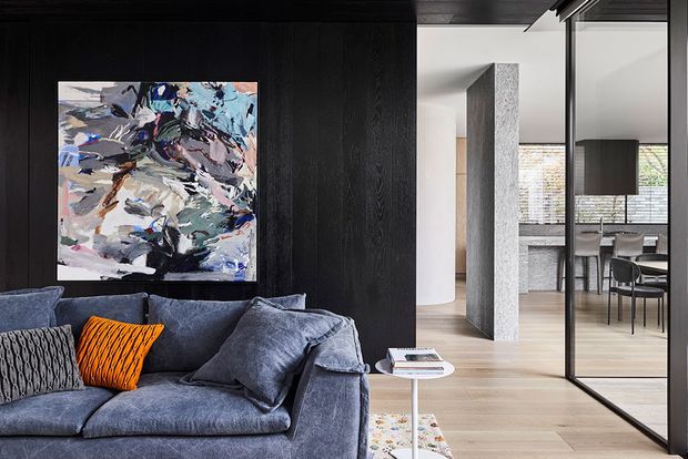 A contemporary open-plan living area with a deep blue-grey sectional sofa and textured cushions positioned against a dramatic black timber feature wall. An abstract artwork provides a colourful focal point, while a slim white side table and patterned rug add lighter accents. Through a black-framed glazed partition, a bright dining area with a timber table and chairs is visible. The space features pale timber flooring, white walls, and a textured grey structural column.