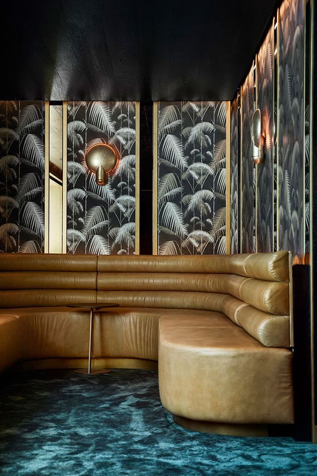 A dramatic, richly layered lounge interior featuring a curved cognac leather banquette wrapping around the room, a rounded upholstered ottoman, dark teal patterned carpet, and black-and-brass detailing. Tropical palm-leaf wallpaper panels line the walls, complemented by sculptural metallic wall lights and a dark ceiling, creating an intimate, theatrical atmosphere.