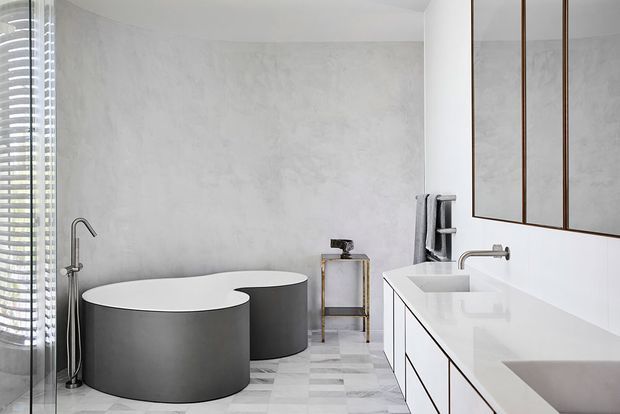 A refined contemporary bathroom featuring two freestanding rounded grey bathtubs, a wall-mounted black-and-white vanity with a white benchtop, a large frameless mirror, a wall-mounted tap, and a pale geometric tiled floor. Textured light grey walls and soft natural light create a calm, minimalist atmosphere.