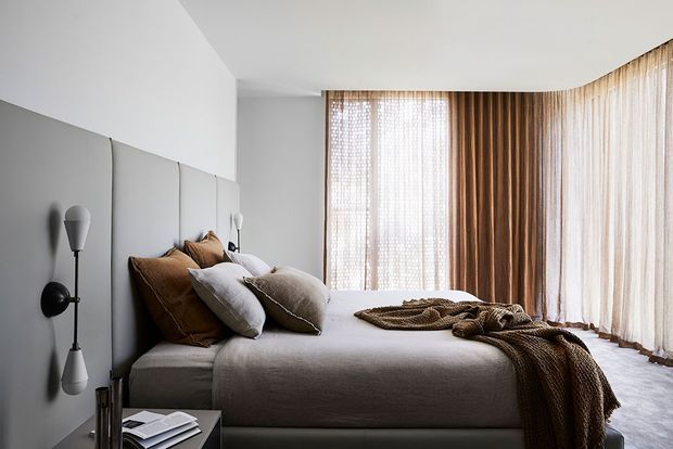 A refined contemporary bedroom with a low upholstered bed, layered neutral bedding, and a sculptural black wall-mounted light. Full-height sheer curtains wrap around the corner windows, creating soft filtered daylight, while warm timber-toned drapery adds texture and contrast. The room has a calm, monochromatic palette with tactile fabrics and minimal furnishings.