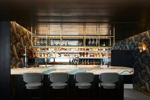 A dramatic, richly detailed home bar with a marble-look counter, four upholstered bar stools, extensive backlit bottle and glass display shelving, dark ribbed cabinetry, dark timber ceiling, patterned wallcovering, and warm metallic lighting.