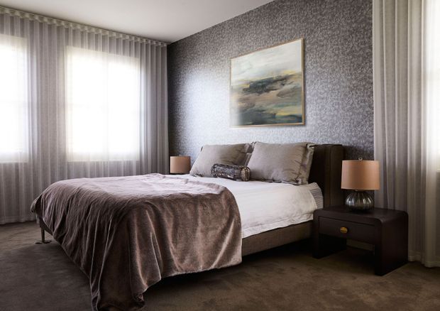 A refined bedroom with a low upholstered bed, layered taupe and grey bedding, dark wood bedside tables, and warm table lamps. Full-height sheer curtains frame bright windows, while a textured dark grey feature wall and framed artwork create a sophisticated, intimate atmosphere. Plush carpet covers the floor.
