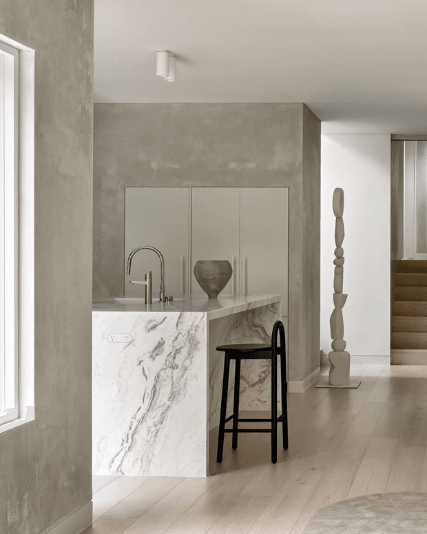 A minimalist open-plan kitchen viewed from an adjoining interior space. The kitchen features a large white marble-look island with an integrated sink and brushed metal tapware, pale built-in cabinetry, and a grey textured plaster or concrete-look wall. Light timber flooring continues through the space, with a black bar stool at the island. A sculptural neutral-toned artwork and timber staircase are visible in the background. The room is softly lit by natural light and a simple ceiling fixture.