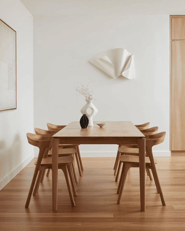 A bright, minimalist dining room with a rectangular natural timber dining table surrounded by matching sculptural wooden chairs. The room has warm timber flooring, white walls, a light timber built-in cabinet at right, simple ceramic tableware and vases, and an abstract white wall sculpture. A large framed artwork is partially visible on the left.