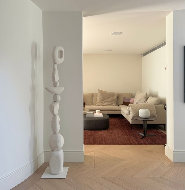 A refined contemporary living room viewed from an adjoining entry or hallway. The room features a large L-shaped beige sectional sofa, dark rounded coffee table, side table, cushions, and a rust-toned area rug. Pale herringbone timber flooring continues through the foreground, with curved white walls, white skirting, recessed ceiling lights, and a linear air-conditioning vent. A sculptural stacked white artwork stands in the entry area.