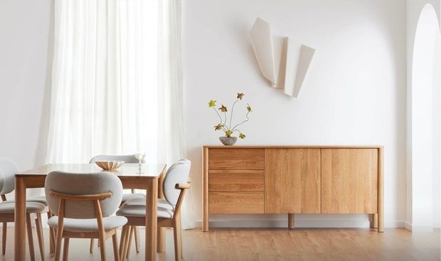 Bright, softly styled dining area with a mid-century-inspired timber dining table and upholstered wooden chairs. A low timber sideboard sits against a white wall beneath sculptural wall lighting, with sheer curtains, pale timber flooring, and an arched opening contributing to the airy, refined appearance.