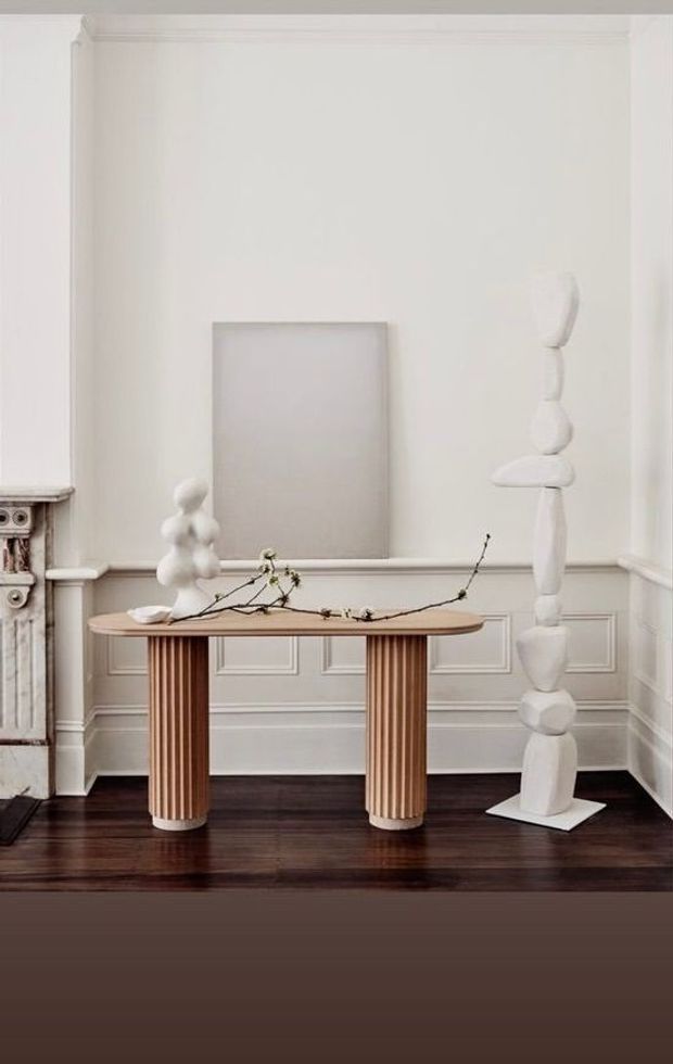 A refined interior vignette featuring a narrow fluted timber console table with rounded edges and cylindrical ribbed legs, styled with minimalist white sculptural objects and a delicate branch. The console sits against a white paneled wall with traditional moulding and wainscoting, above a dark polished timber floor. A partial ornate fireplace or mantel is visible at the left edge. The composition is elegant, monochromatic, and gallery-like.