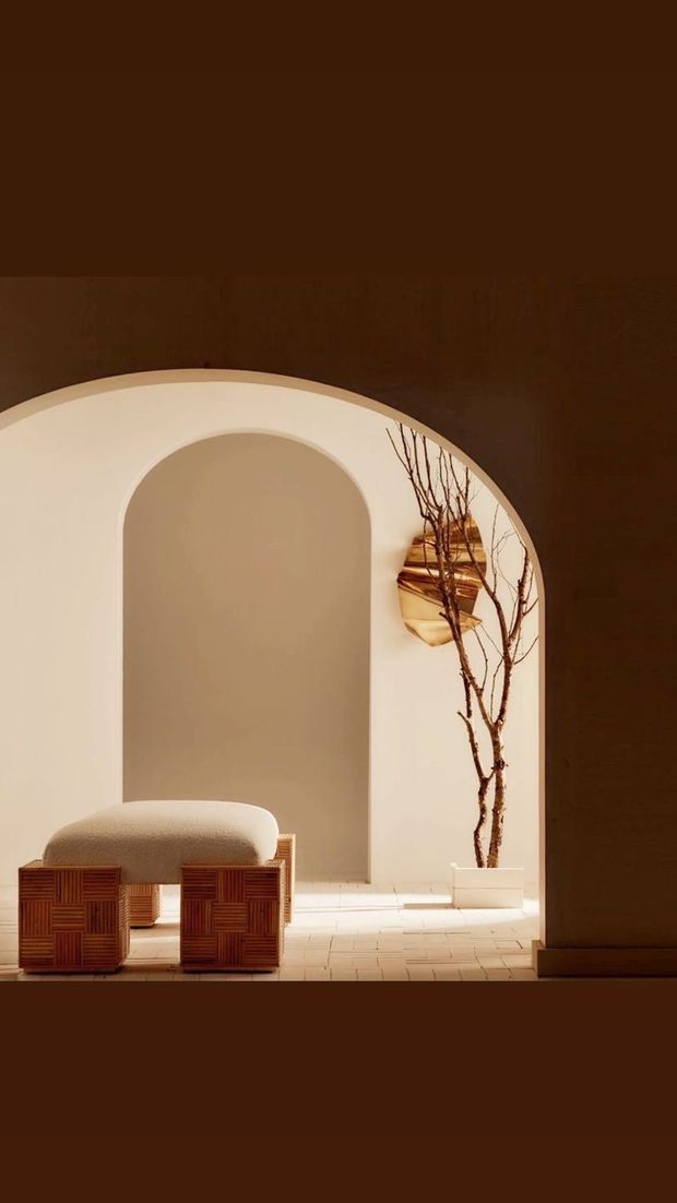 A warm, sculptural interior featuring a large rounded archway with a smaller recessed arch, creamy plaster walls, and pale tiled flooring. A low upholstered bench sits on a richly patterned timber base, while a tall branch arrangement and sculptural wall decor add natural and artistic accents. The space is softly lit with a minimalist, refined atmosphere.