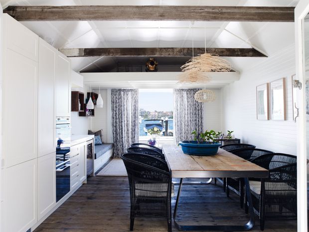 Bright coastal-style open-plan dining area with a long timber dining table and black woven chairs, white built-in cabinetry and kitchen joinery, dark timber flooring, exposed rustic ceiling beams, and woven pendant lights. Large glazed doors at the far end open toward an outdoor view.