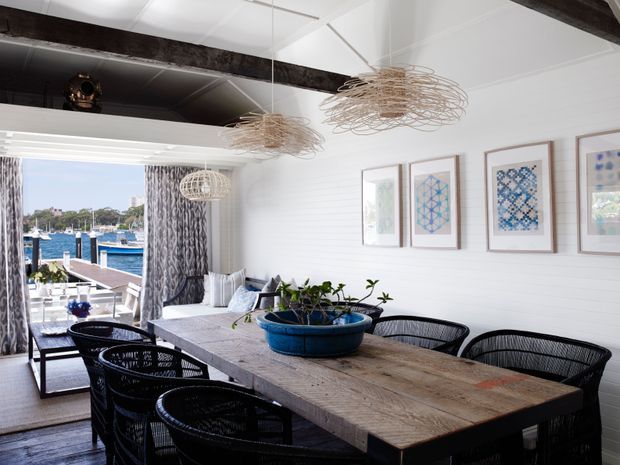 Bright coastal dining room with a long rustic timber dining table, black woven dining chairs, white horizontal wall panelling, exposed dark ceiling beams, and sculptural woven pendant lights. Framed geometric artwork lines the wall, while large glazed doors open to a waterside setting with boats and a marina visible beyond.