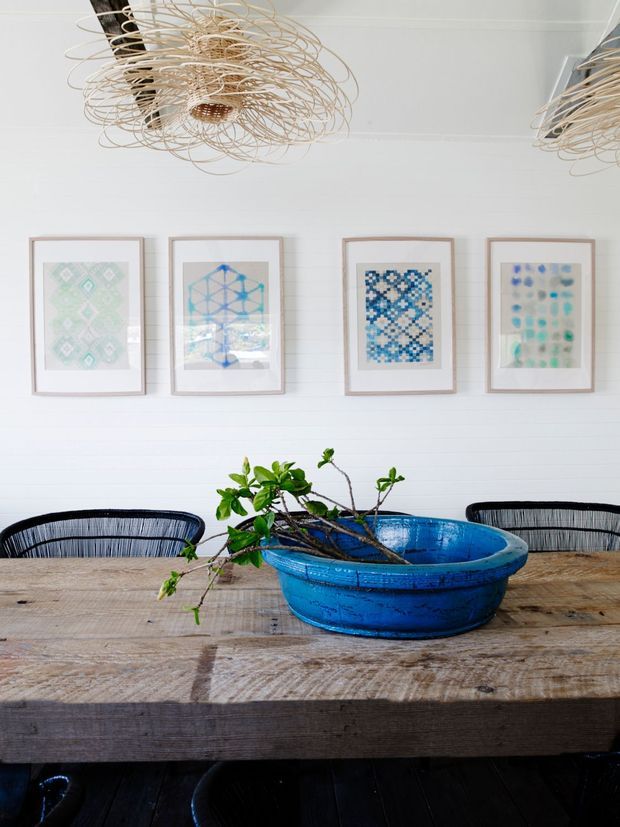 Bright dining area with a rustic timber dining table, black woven-back dining chairs, a vivid blue planter holding leafy branches, framed geometric artwork, and sculptural woven pendant lights. White horizontal wall panelling and dark timber flooring create a relaxed coastal, eclectic feel.