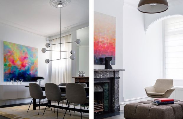 A refined interior showing a dining area and adjacent sitting nook. The dining space features a long table with upholstered charcoal-grey chairs, a contemporary sculptural pendant light, white walls, sheer curtains, and a large abstract artwork. The adjoining sitting area includes a brown upholstered lounge chair and ottoman, a dark ornate fireplace surround, wall art, and plantation shutters. The overall scheme combines classic architectural detailing with contemporary furnishings and colourful artwork.
