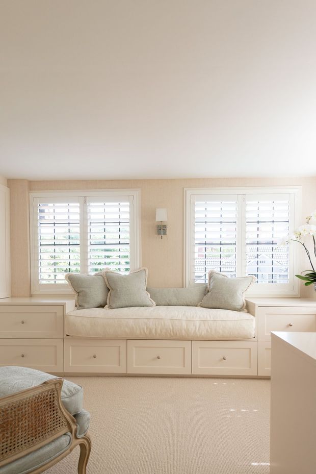 Bright, softly styled sitting room with a built-in window seat and drawers beneath two sets of white plantation-shutter windows. The room has warm beige textured wallcovering, cream upholstery and carpet, white built-in cabinetry, a delicate wall sconce, and a partially visible wicker-framed armchair and houseplant. Natural daylight creates an airy, calm atmosphere.