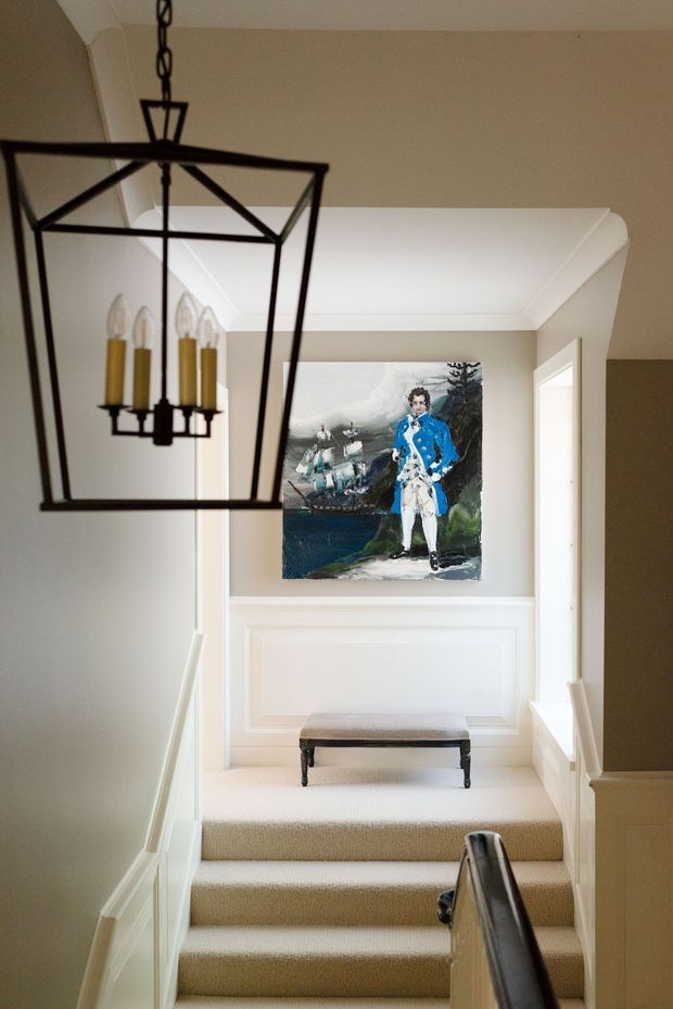 A refined residential stairway with carpeted steps, white painted paneled walls and moulding, and a dark timber handrail. A large black metal lantern pendant hangs in the foreground, while a bench and colourful portrait artwork decorate the upper landing.