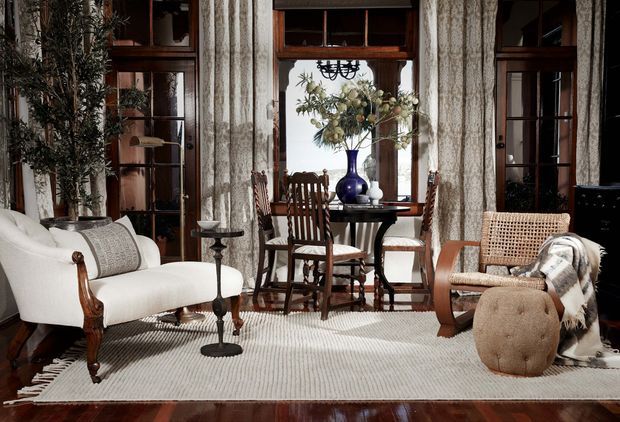 A refined, traditional sitting room with a small dining or occasional table centered before tall timber-framed glazed doors and windows. The room features a white upholstered settee with carved dark-wood arms, a pair of dark timber chairs, a woven armchair, a tufted round ottoman, a large light textured rug, patterned full-height curtains, and a decorative floral arrangement in a deep blue vase. The space has layered natural textures, rich timber detailing, and abundant filtered daylight.