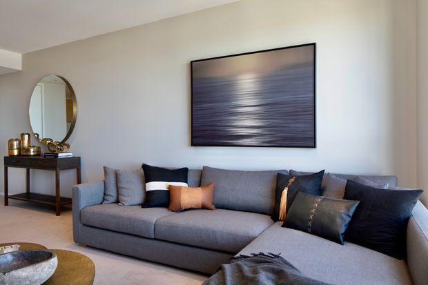 A refined living room with a large grey L-shaped sectional sofa, layered dark and neutral cushions, a slim dark wood console table with an oval mirror and decorative objects, and a large framed abstract artwork above the sofa. The room has pale neutral walls, soft carpeted flooring, and warm natural light.