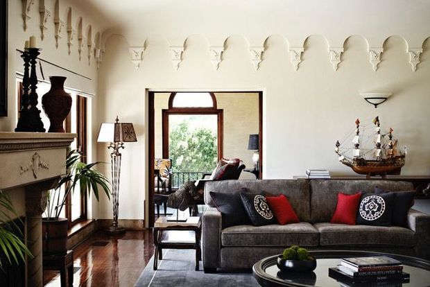 Elegant traditional living room with a dark upholstered sofa, patterned accent cushions, polished timber flooring, decorative fireplace mantel, arched doorway, ornate wall detailing, and classic furnishings. A balcony or exterior garden is visible through the open doorway.