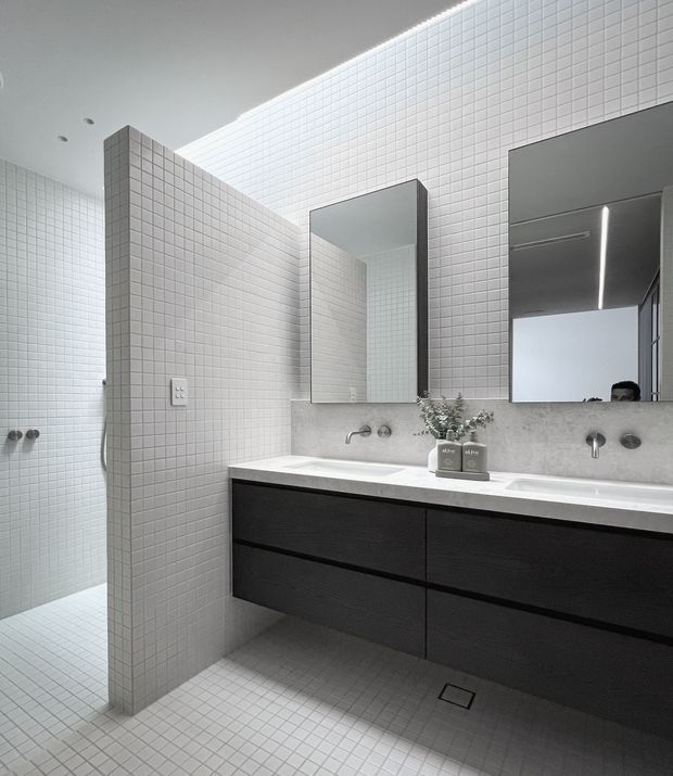 Contemporary bathroom with a large floating double vanity, dark wood-look drawers, white benchtop, wall-mounted taps, twin rectangular mirrors, and extensive small-format white square tiling across the walls and floor. A tiled shower area is partially screened by a tiled partition wall. The space has integrated linear ceiling lighting and a restrained monochromatic palette.