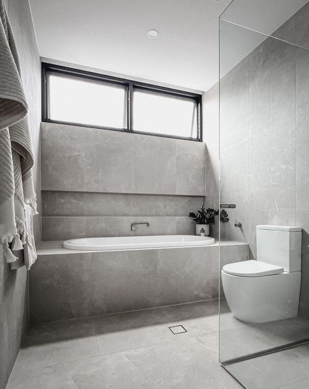 A compact contemporary bathroom in a neutral grey palette, featuring a built-in freestanding-style bathtub beneath high horizontal windows, a wall-mounted tap, large-format tiled walls and floor, a frameless glass shower screen, and a white toilet. A small plant and textured towels add subtle warmth.