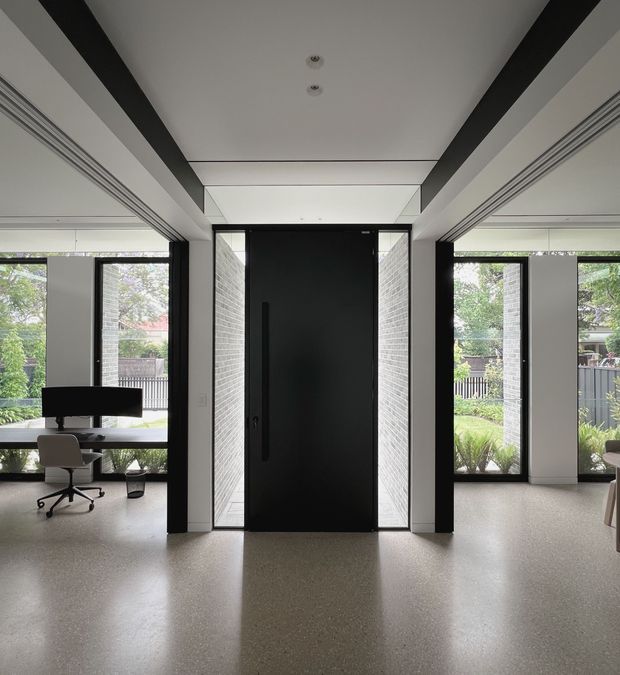 A minimalist residential entryway centered on a tall matte-black pivot door with narrow glazed sidelights and textured light brick reveals. Black structural or trim elements frame the opening, while expansive windows on both sides overlook a landscaped outdoor area. The space has a light terrazzo-like floor, white walls and ceiling, integrated linear ceiling detailing, and a small home office visible to the left.