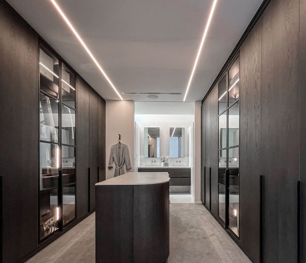 A refined, contemporary dressing room or walk-in wardrobe with full-height dark timber cabinetry and black-framed glazed display doors on both sides. A central island with a pale stone benchtop anchors the room, while a bathroom vanity and mirrors are visible at the far end. Linear recessed LED lighting runs along the ceiling, and the floor has a smooth grey concrete-like finish.